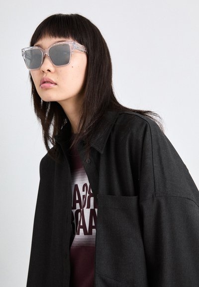 Lunettes de soleil claires avec une monture angulaire et translucide. Chemise noire superposée sur un t-shirt graphique bordeaux. Texture douce, motifs subtils visibles.