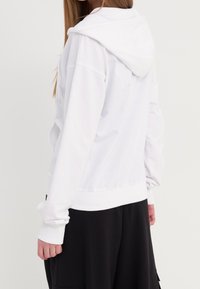 Sweat à capuche blanc avec une coupe décontractée, doté d'une capuche à cordon, de poignets et d'un ourlet côtelés, associé à un pantalon noir ample. Texture de tissu lisse.