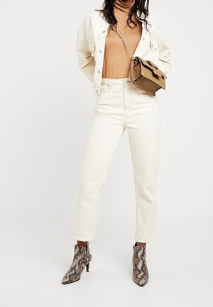 Femme portant un col roulé beige, une veste en denim crème et un pantalon, des bottines à imprimé serpent et un petit sac marron en bandoulière.