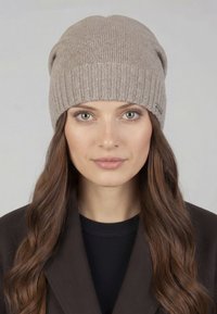 Gebreide muts in licht beige met een geribbelde structuur, voorzien van een omgeslagen boord en een klein metalen accent aan de zijkant. Zachte textuur.