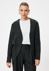 Cardigan grigio scuro dal taglio ampio, con bordi a coste, apertura frontale e maniche lunghe, indossato su una camicia bianca e pantaloni neri su misura.