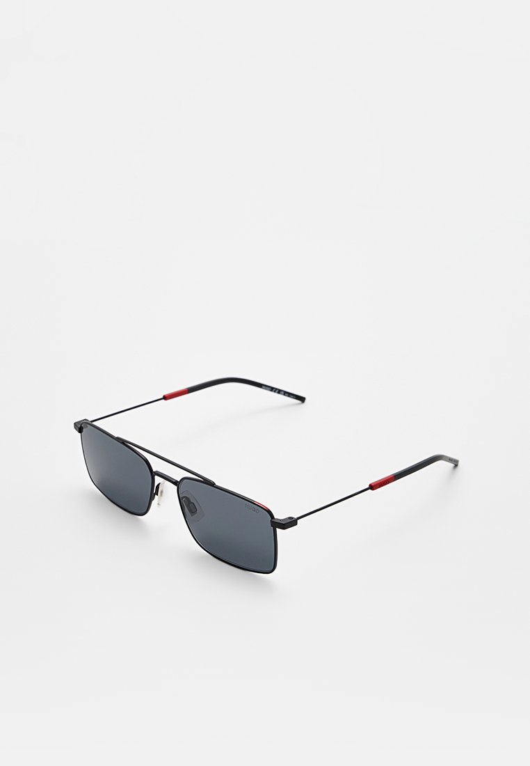 Schwarze rechteckige Sonnenbrille mit dünnem Metallrahmen und roten Akzenten an den Bügeln, auf einer weißen Oberfläche präsentiert.