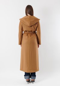 Cappotto lungo marrone con un grande cappuccio, vita cintata e taglio dritto. Presenta una superficie morbida e liscia ed è abbinato a jeans risvoltati e tacchi.