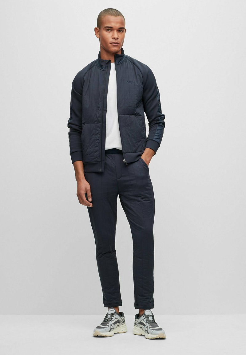 Homme portant une veste de survêtements matelassée bleu marine et un pantalon assorti sur un T-shirt blanc, associé à des baskets grises et blanches, se tenant devant un fond uni.