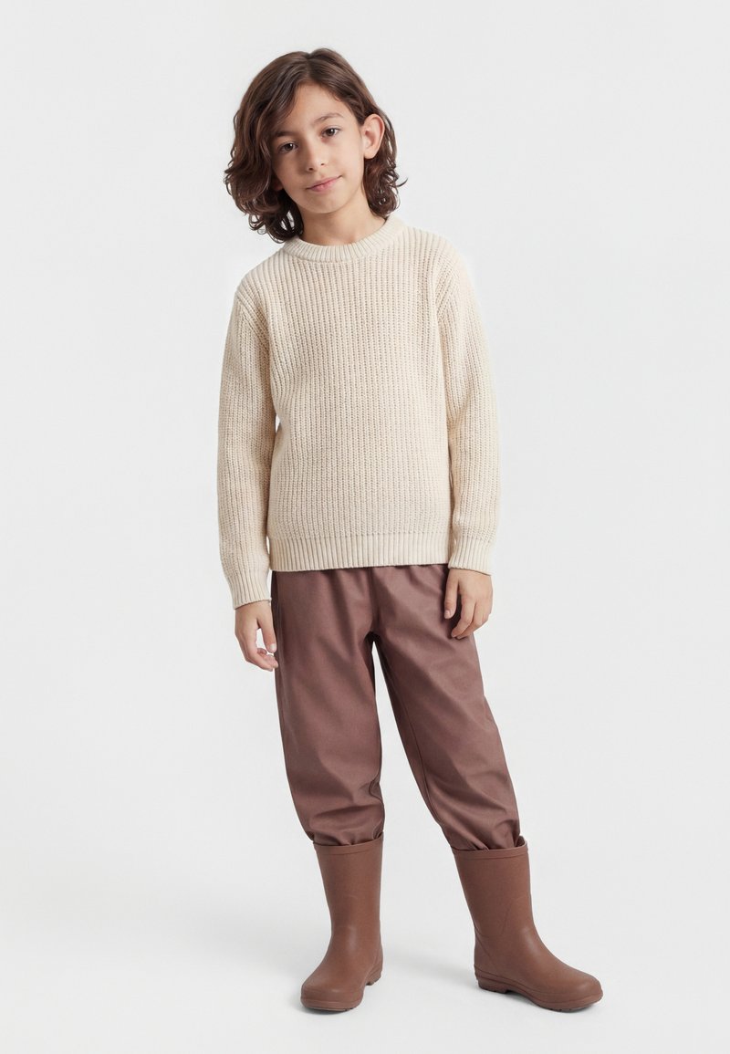 Copil cu păr castaniu până la umeri, purtând un pulover crem tricotat, pantaloni maro băgați în cizme maro asortate, stând pe fundal alb.