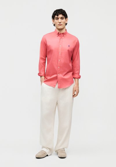 Νεαρός άνδρας στέκεται με τα χέρια στις τσέπες, φορώντας ένα κοραλί πουκάμισο με κουμπιά, λευκό παντελόνι φαρδύ και μπεζ μοκασίνια σε απλό φόντο.