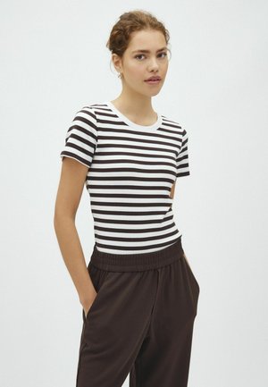 Tânără purtând un tricou cu mâneci scurte, alb-negru în dungi și pantaloni închiși la culoare, cu mâinile în buzunare, stând în fața unui fundal alb simplu.
