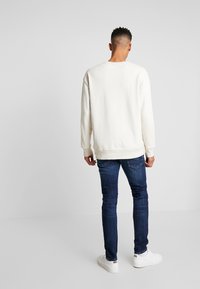 Joven de pie de espaldas, vestido con una sudadera blanca, jeans azules y zapatillas blancas sobre un fondo liso de color gris claro.