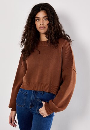 Brown knitted sweater with a cropped design, ribbed cuffs, and round neckline, paired with blue jeans. Textured surface detail visible.