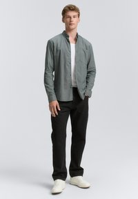 Lys grøn skjorte med knaplukning og brystlomme, parret med sorte bukser og hvide sneakers. Stoffet ser glat og tekstureret ud.