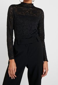 Schwarzes Spitzenoberteil mit langen Ärmeln und hohem Kragen, mit Blumenmustern und transparenter Textur, kombiniert mit hoch taillierten schwarzen Hosen.