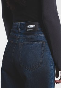 Vaqueros de mezclilla azul oscuro con cintura alta, con una etiqueta negra de marca en la parte posterior, costuras reforzadas y bolsillos clásicos.