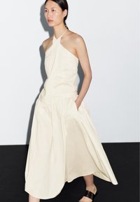 Female model wearing a sleeveless, cream-colored halter dress with pockets, walking on a black and white background.