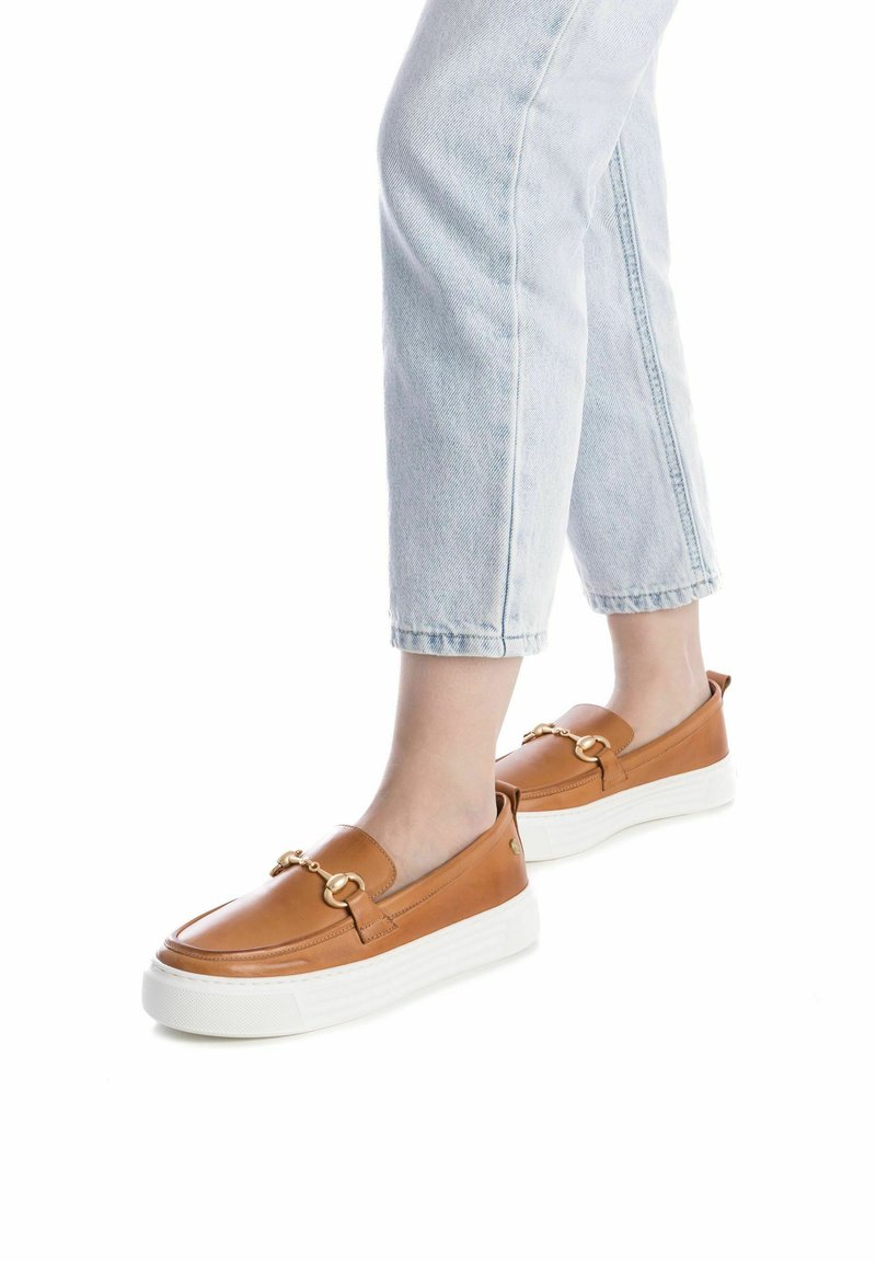 Zapatos slip-on de cuero marrón con suelas de plataforma blanca y detalles de hardware dorado. Textura suave, forma redondeada y diseño minimalista.