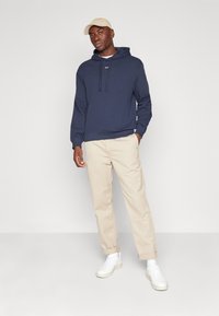 Felpa navy con coulisse e logo, abbinata a pantaloni beige a sigaretta e sneakers bianche, su uno sfondo bianco semplice.