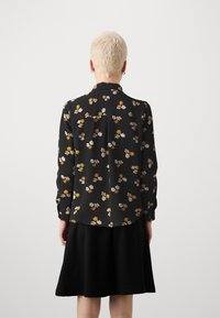 Blusa floral preta com padrões coloridos, apresentando mangas longas, um yoke nas costas e uma bainha curvada. Usada com uma saia preta simples.