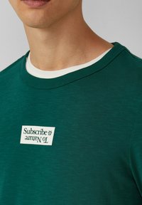 Zielony, okrągły sweter z teksturowanej tkaniny z białym wewnętrznym kołnierzykiem. Posiada białą metkę z napisem "Subskrybuj Naturę".