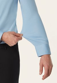 Lichtblauwe langermige shirt met een gladde textuur; een persoon trekt voorzichtig aan de manchet. Zwarte broek is gedeeltelijk zichtbaar.