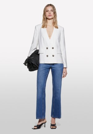 Blazer blanc à double boutonnage avec des boutons métalliques, associé à un jean bleu, des pantalons évasés, et des escarpins noirs à bout ouvert.