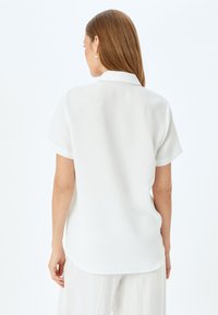 Blusa bianca a maniche corte con colletto, caratterizzata da una vestibilità comoda e una texture liscia. Il retro ha un orlo arrotondato e dettagli minimi.