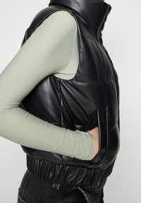 Osoba ubrana w czarną skórzaną kamizelkę puchową z wysokim kołnierzem na jasnej zielonej bluzce z długim rękawem, z ręką w kieszeni kamizelki, w połączeniu z czarnymi dżinsami.