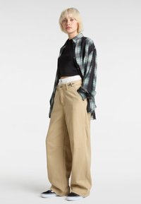 Schwarzes Croptop unter einem grün-schwarz karierten Hemd, kombiniert mit weiten beigen Hosen und schwarzen Schuhen mit weißen Sohlen.
