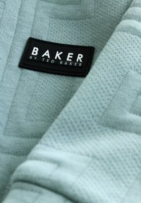 Lyseblåt tekstureret stof med et geometrisk mønster. Indeholder et sort mærke med teksten "BAKER by TED BAKER" i hvidt.