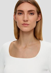 Witte geribde top met een vierkante halslijn. Het model heeft steil blond haar, minimale make-up en kleine gouden hoepel oorbellen.