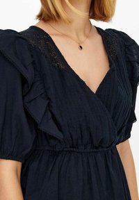 Abito blu navy con maniche corte a volant, corpino plissettato e dettagli in pizzo lungo il collo. Tessuto morbido con un design fluido.