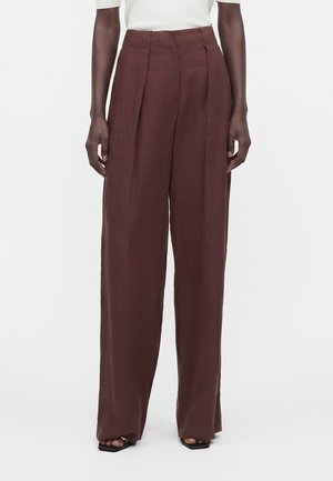 Persoon die een hoge taille wijde bruine broek en een wit topje draagt, staat met ontspannen handen langs het lichaam tegen een witte achtergrond.