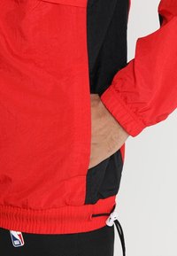 Giacca rossa e nera con una finitura strutturata, polsini elasticizzati e orlo regolabile. Presenta una tasca laterale e il logo NBA nella parte inferiore.