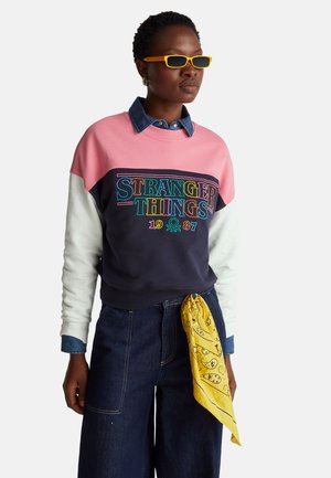 Vrouw draagt een Stranger Things sweatshirt in roze, marineblauw en wit, een spijkershirt, donkere jeans met een gele bandana en oranje zonnebril.