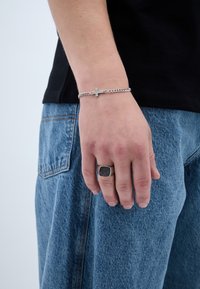 Stříbrný řetízkový náramek s křížkovým detailem na zápěstí, nošený s černou košilí a volnými modrými džínami. Velký stříbrný prsten na ruce.