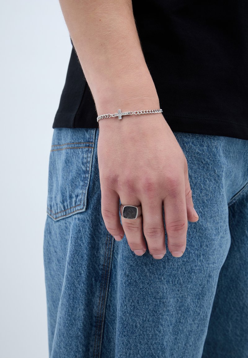 Stříbrný řetízkový náramek s křížkovým detailem na zápěstí, nošený s černou košilí a volnými modrými džínami. Velký stříbrný prsten na ruce.