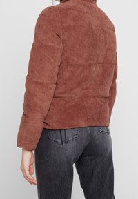 Hnědá manšestrová péřová bunda s vertikálními pruhy, vysokým límcem a elastickými manžetami, kombinována s tmavými džíny.