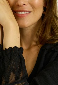 Haut noir texturé avec des détails en dentelle aux poignets, laissant entrevoir la peau en dessous. Le décolleté est plongeant et le tissu est léger.