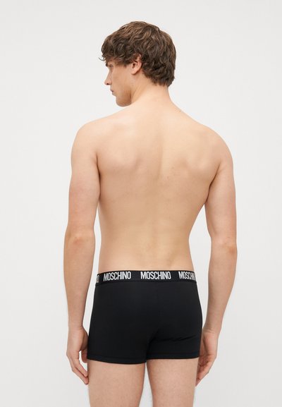 Must musta lühikesed püksikud valge Moschino logo elastse vööga, istuv disain ja sujuv tekstuur, nähtavad seljalt palja torso peal.