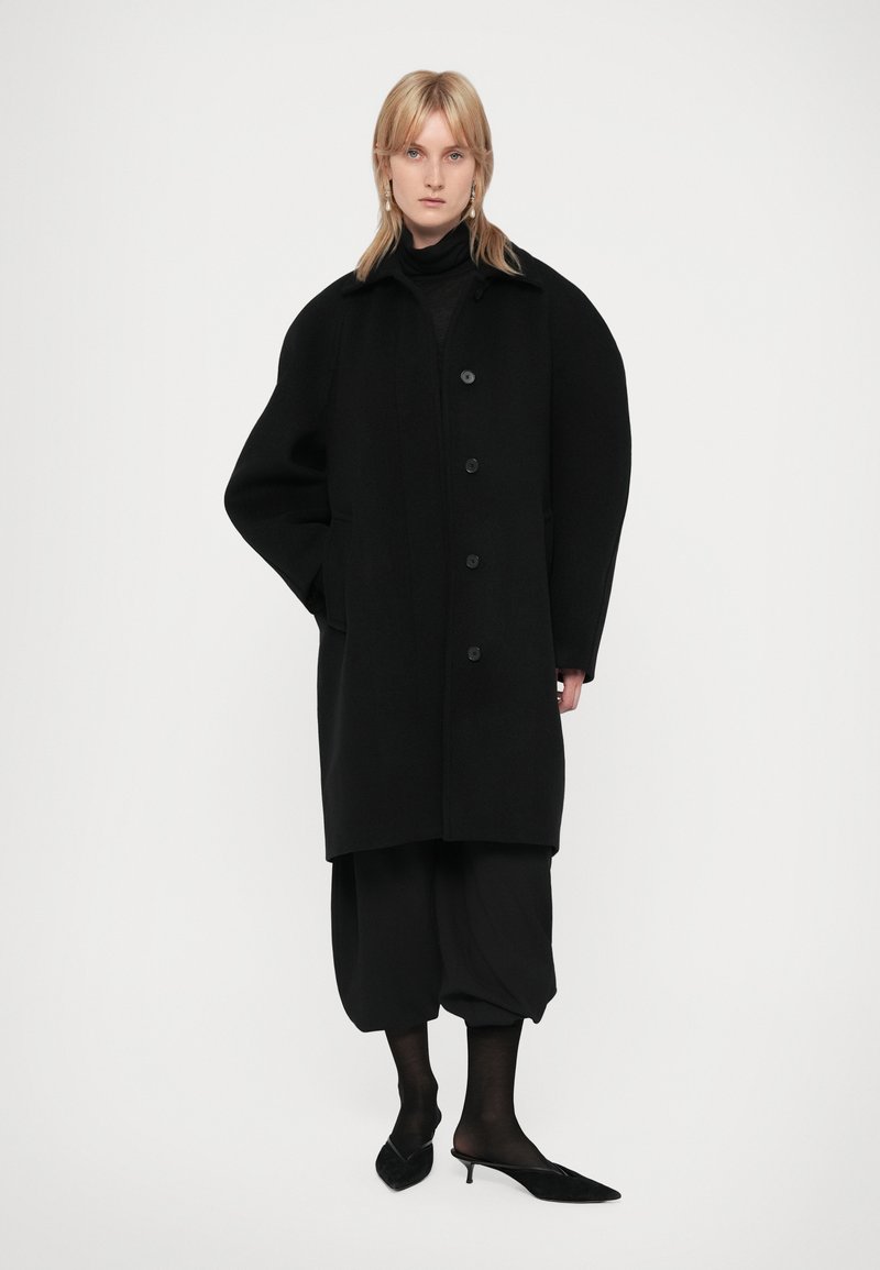 Svart ullrock med stora axlar och knäppning fram, kombinerad med lösa svarta byxor och spetsiga svarta klackskor.