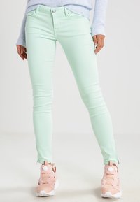 Un jean skinny vert menthe présente une texture lisse et un détail de fermeture éclair à la cheville, associé à des chaussures de sport roses pour le contraste.