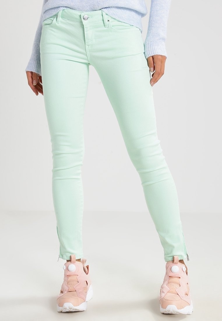 Un jean skinny vert menthe présente une texture lisse et un détail de fermeture éclair à la cheville, associé à des chaussures de sport roses pour le contraste.
