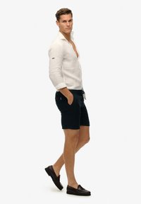 Weißes Leinenhemd mit Knöpfen, blaue Shorts mit Taschen, kombiniert mit dunklen Slippers. Hat eine lockere Passform und hochgekrempelte Ärmel.