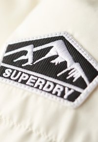 Patch ricamato in bianco e nero con design di montagna e testo "SUPERDRY" su uno sfondo in tessuto color crema.