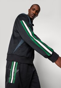 Veste de sport noire avec des accents rayés verts, blancs et verts. Comprend un col zippé et des poignets côtelés ; portée avec un short noir assorti.