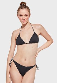 Bikini negro con top triangular y parte inferior ajustable, presentado con tirantes estampados en negro, blanco y rojo. Textura suave, diseño minimalista.