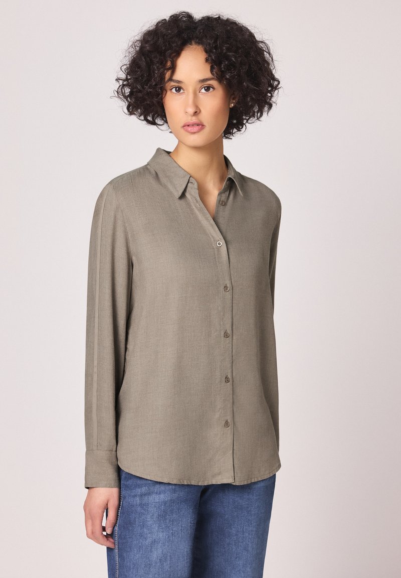 Femme aux cheveux bouclés portant un chemisier taupe à manches longues et boutonné, ainsi qu'un jean bleu, debout devant un fond uni.