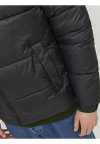 Sort pufferjakke med quiltet tekstur, der har en sidelomme, lynlås og grøn ribbet kant. Materialet fremstår glat og skinnende.