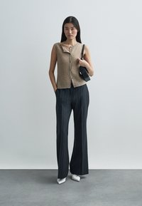 Beige ärmelloses Strickoberteil mit Knopfleiste, kombiniert mit dunklen, Nadelstreifen-weiten Hosen und weißen spitzen Absätzen, hält eine schwarze Handtasche.