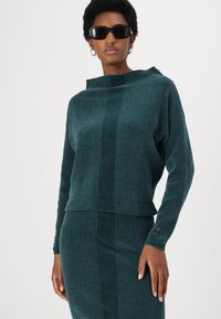 Turkoois gebreide trui met een hoge halslijn en lange mouwen, voorzien van een geribbelde zoom en contrasterende textuurpanelen. Vergezeld van een bijpassende rok.