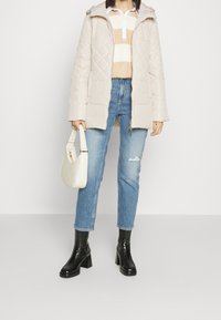 Vrouw in een beige gewatteerde jas, gestreept beige en wit sweater, gescheurde blauwe jeans, zwarte hoge hakken, met een witte handtas.