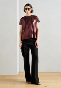 Metalický burgundský top s krátkymi rukávmi, doplnený o čierne široké nohavice. Model drží čiernu kabelku a má na sebe oversized slnečné okuliare.